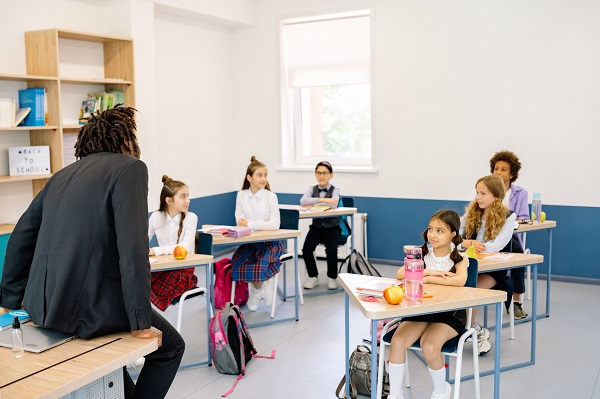 Six students sitting on the school tables, each having an apple and a water bottle on it, a teacher leaning against the table.