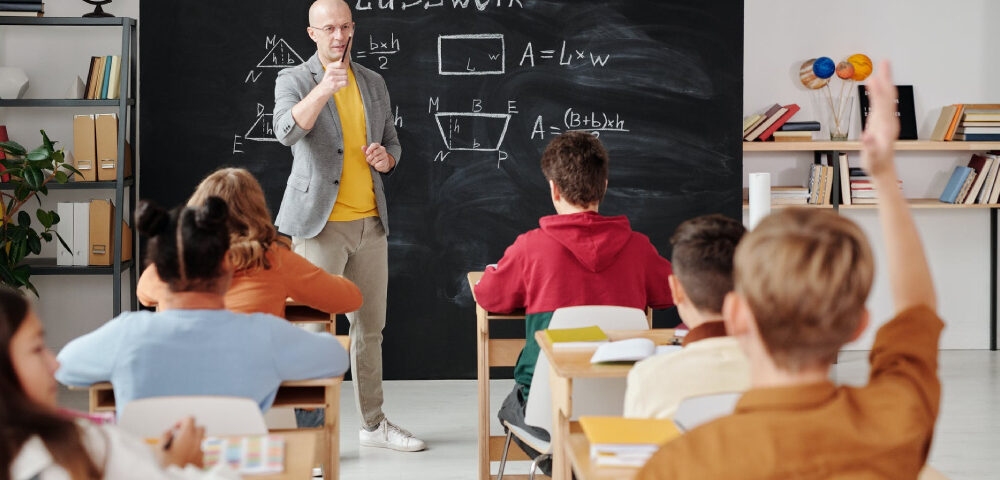 A school teaching is teaching his students a lesson, while one student in the last row, puts his hands up.