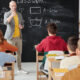 A school teaching is teaching his students a lesson, while one student in the last row, puts his hands up.