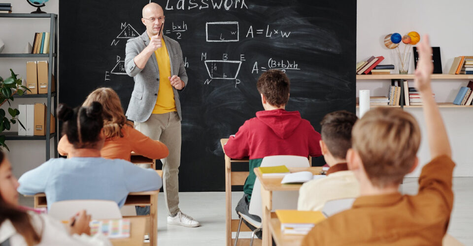 A school teaching is teaching his students a lesson, while one student in the last row, puts his hands up.