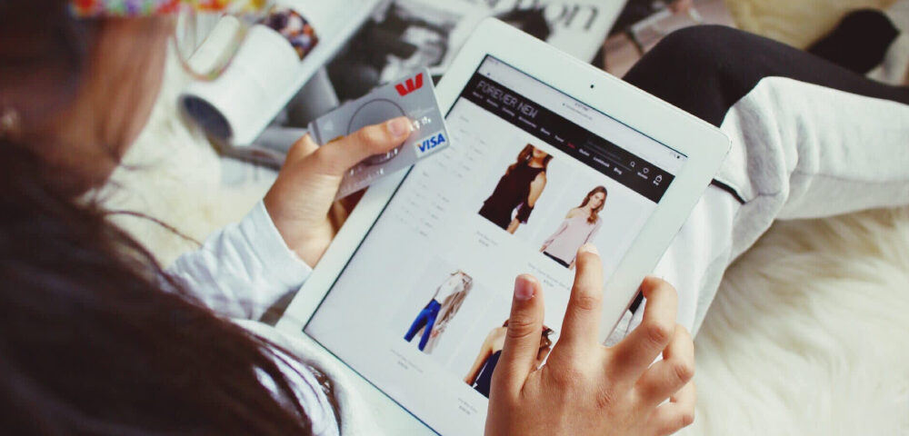 A girl is shopping apparels on the ecommerce platform, keeping her tablet on her lap and having debit card in her hand.