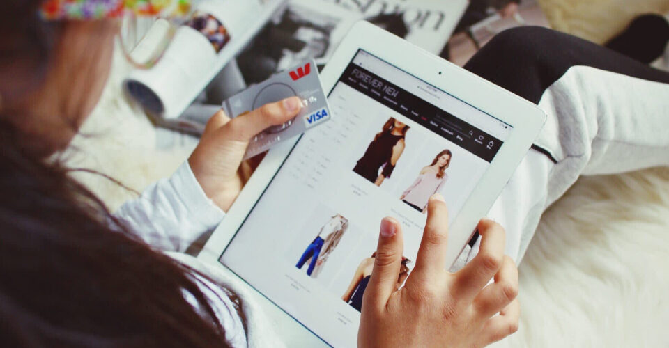 A girl is shopping apparels on the ecommerce platform, keeping her tablet on her lap and having debit card in her hand.